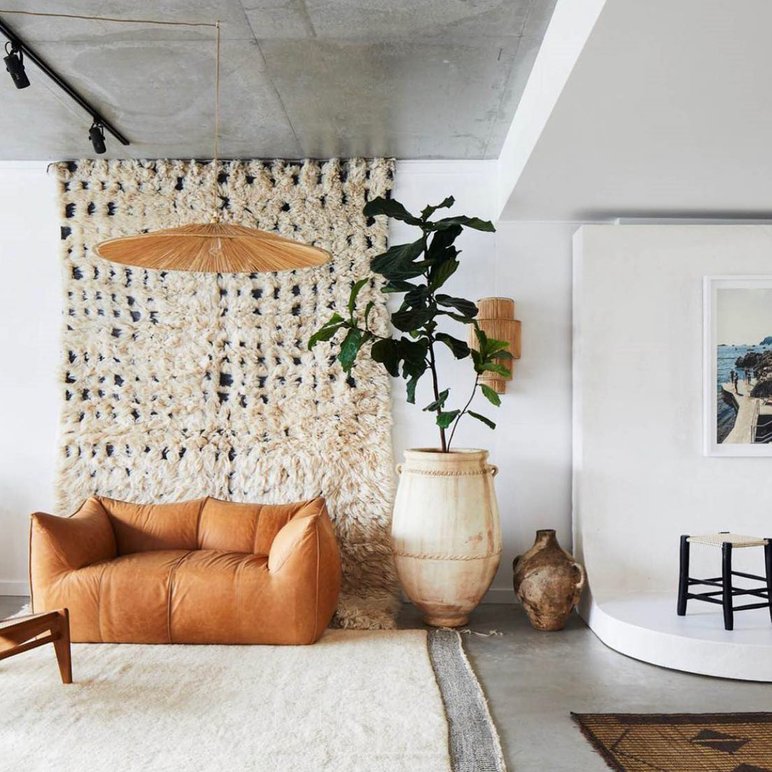 Salon au style bohème avec canapé en cuir caramel, grand tapis texturé, tenture murale en laine, plante verte dans un vase en terre cuite et éléments décoratifs de Cœur d’Artichaut