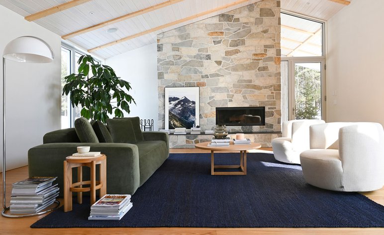 Salon moderne et chaleureux avec mobilier design et cheminée en pierre de chez Élément de Base Salon lumineux au style contemporain avec un canapé vert en velours, fauteuils blancs arrondis, tapis bleu profond et cheminée en pierre naturelle, créant une ambiance chaleureuse et élégante de chez Élément de Base