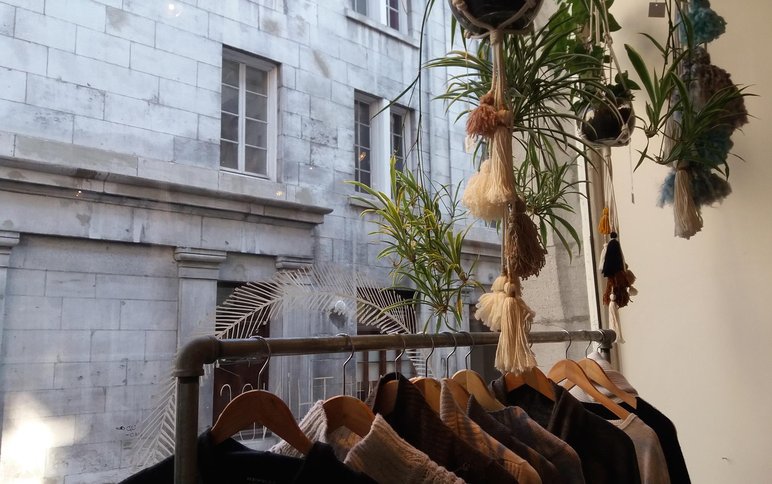 Vêtements suspendus sur cintres près d'une fenêtre avec des plantes suspendues en macramé, vue sur un bâtiment en pierre à l'extérieur Par Maison Pépin
