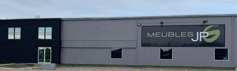 Façade extérieure de l'Entreprise Meubles JPG à Dolbeau-Mistassini, avec enseigne visible sur un grand bâtiment gris et stationnement à l'avant.