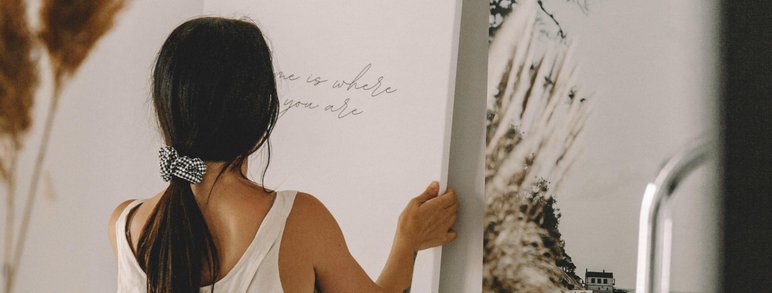 Femme vue de dos tenant une toile décorative sur laquelle est écrit 'Home is where you are', dans un intérieur doux et naturel, aux accents bohèmes et apaisants par Oleka Canvas
