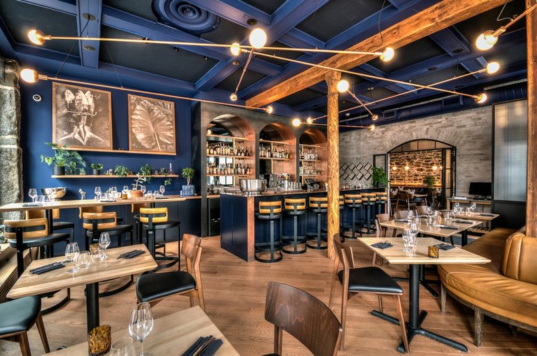 Intérieur d’un restaurant moderne au design élégant avec bar central par Taktik Design Intérieur d’un restaurant moderne au design élégant avec bar central, murs en pierre et plafond bleu marine, tables en bois et éclairage suspendu par Taktik Design