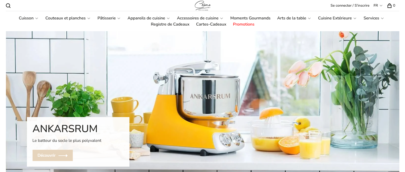 Mélangeur sur socle Ankarsrum jaune sur un comptoir de cuisine lumineux, entouré de verres de jus d'orange, d'herbes fraîches, d'ustensiles en bois et d'un bouquet de tulipes chez Boutique Crème.