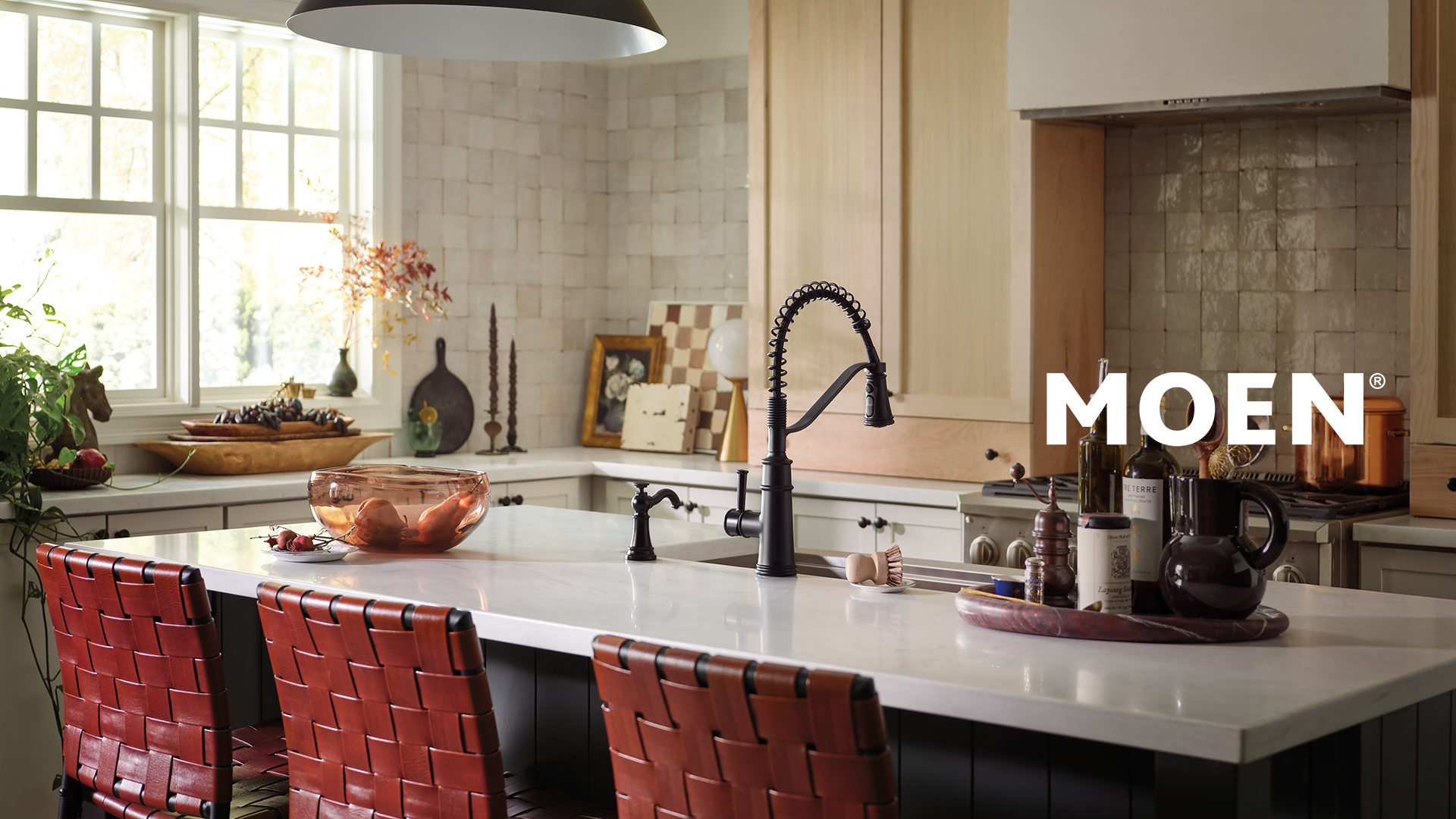 Cuisine moderne avec un robinet noir de style professionnel MOEN installé sur un îlot central en marbre blanc, entouré de chaises en cuir tressé rouge, avec le logo MOEN affiché en blanc au premier plan Cuisine moderne avec un robinet noir de style professionnel MOEN installé sur un îlot central en marbre blanc, entouré de chaises en cuir tressé rouge, avec le logo MOEN affiché en blanc au premier plan